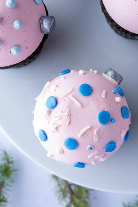 a pastel pink christmas ornament cupcake with vegan blue and white sprinkles.