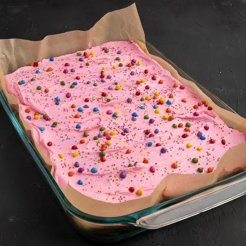 a chocolate cake with pink frosting and rainbow sprinkles in a lasagna pan lined with parchment paper.