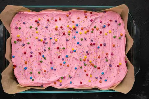 a chocolate cake with pink frosting and rainbow sprinkles in a lasagna pan lined with parchment paper.