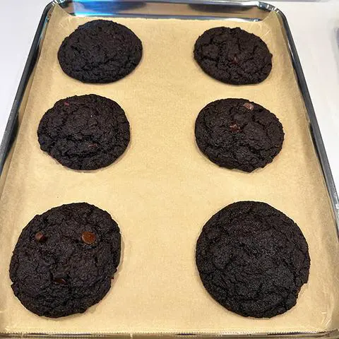 6 baked chocolate cookies on a baking sheet covered in unbleached parchment paper.