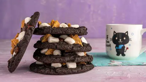 stack of vegan chocolate cookies with one leaning on the stack showing the top of it and a cup with a black cat on it to the right of the stack.